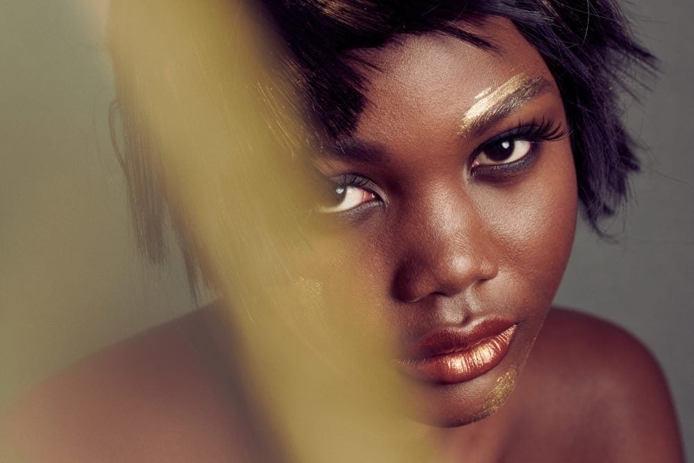 portrait of model with dramatic gold and bronze makeup and her face partially obscured with golden organza